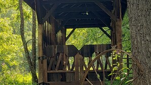 Ein rustikales Holzgebäude mit geneigtem Dach und einer Radmechanik, umgeben von üppigem Grün und Bäumen.