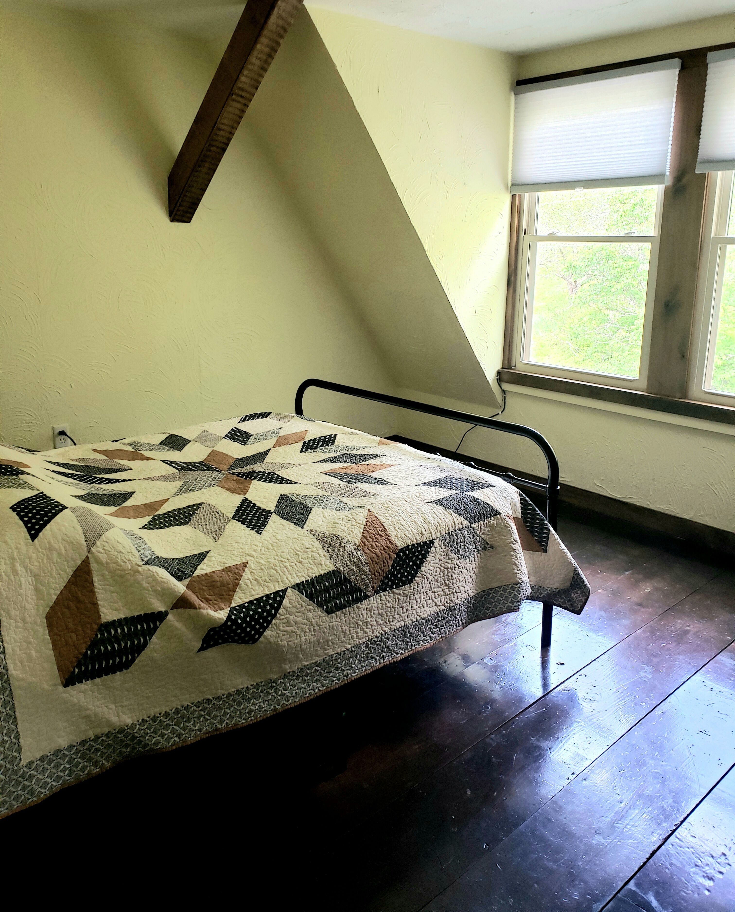 Une petite chambre avec un lit simple, un plafond en pente, une fenêtre avec des stores et une courtepointe sur le lit.