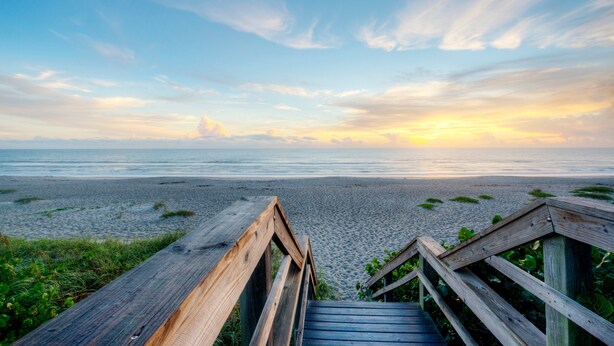 Una pasarela de madera que lleva a una playa, con vista despejada al océano y un atardecer de fondo.