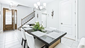 A modern dining area with a glass-top table, white chairs, a chandelier, and a staircase leading to another room. - Family Fun Beach Townhome w/ Pool, BBQ & Game Room! 🌊🏖️ (Indian Harbour Beach)