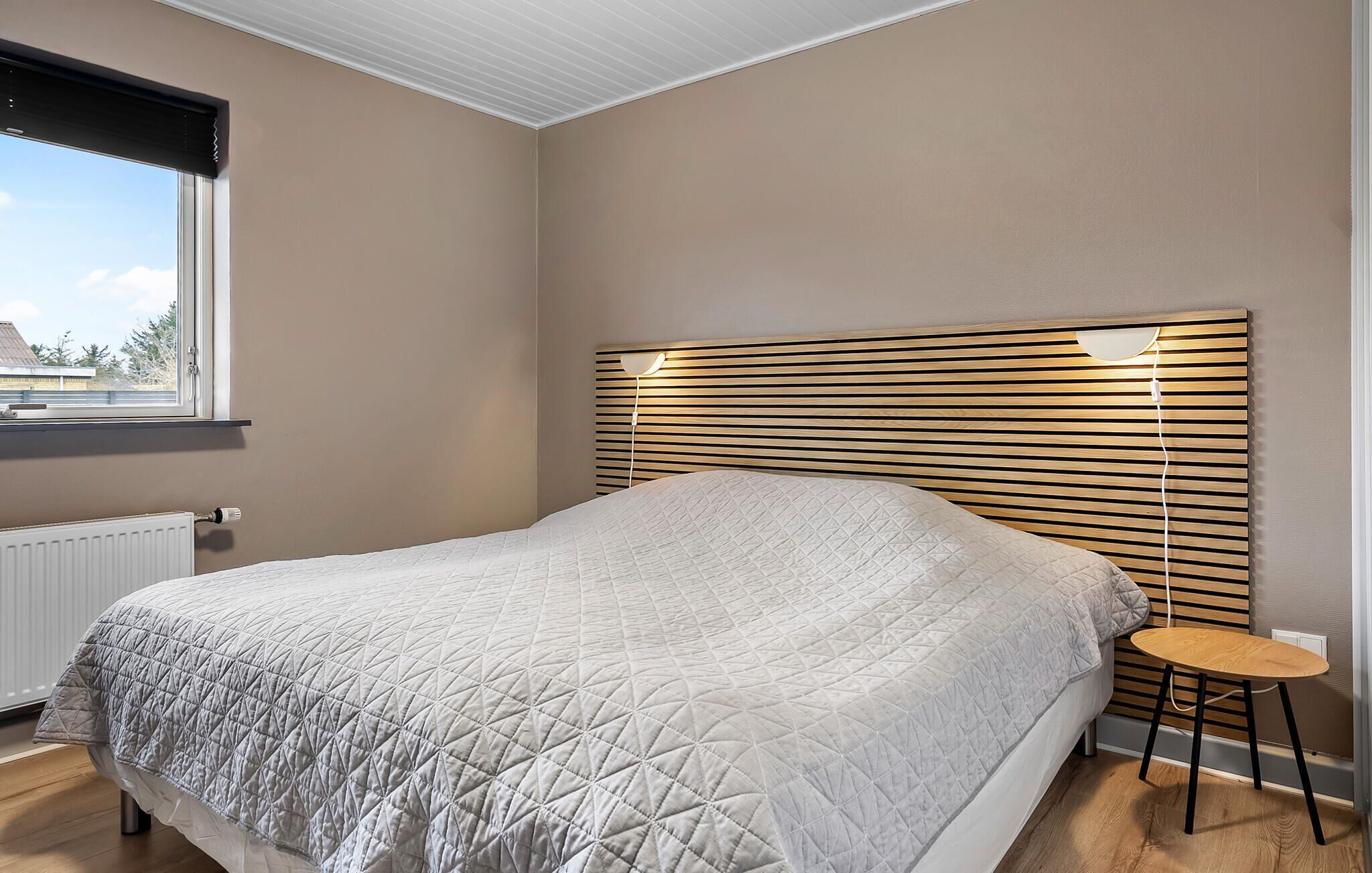 Quarto com cama, janela, radiador, mesa de cabeceira e abajur.