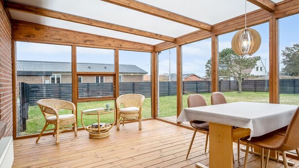 A covered patio with wooden flooring, wicker chairs, a table, and large windows offering a view of a backyard and neighboring houses. - Amazing home in Vestervig with WiFi (Vestervig)