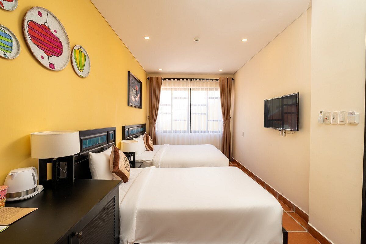 Una camera d'albergo con due letti, una scrivania con lampada, una televisione e una finestra con tende.