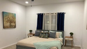 A bedroom with a bed, two nightstands, a window with curtains, a painting on the wall, and a carpeted floor.
