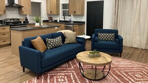 A modern kitchen with wooden cabinets, a black range hood, and a central island. There is a blue sofa with patterned cushions, a round wooden coffee table, and a small side table with a plant.