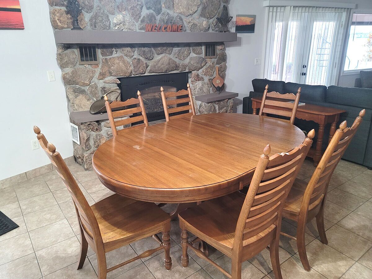 Une table de salle à manger ronde en bois, entourée de six chaises, une cheminée en pierre et une pancarte « BIENVENUE » placée au-dessus.