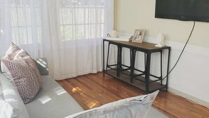 Quarto com piso de madeira, sofá cinza com almofadas estampadas, uma pequena mesa preta com um objeto branco sobre ela e uma TV fixada na parede.