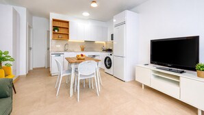 Een kleine keuken met een wasmachine, een eettafel met stoelen, een tv op een meubel en een plant.