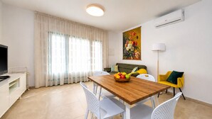 Een moderne woonkamer met een houten eettafel, witte stoelen, een bank, een televisie en een schilderij aan de muur.