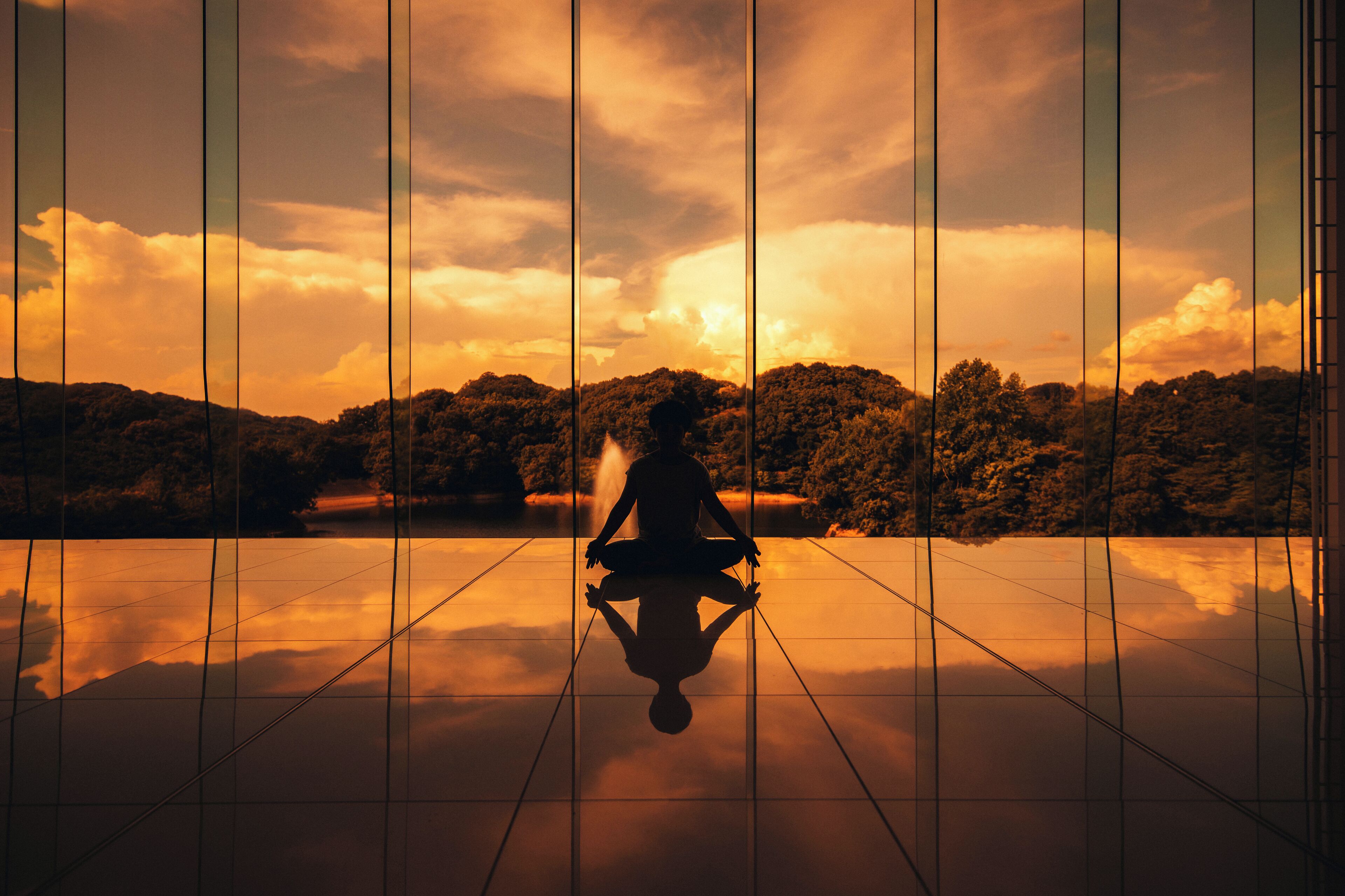 A person meditating in a room with a view of a sunset over a forested landscape.