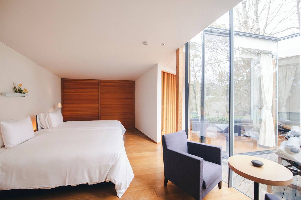 Une chambre moderne dotée d’un grand lit, d’une tête de lit en bois, d’un coin salon avec une table et d’un mur en verre offrant une vue sur l’extérieur.
