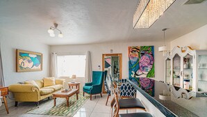 Un salon comprenant un canapé, des fauteuils, une table basse et un téléviseur. Il y a un tableau accroché au mur et une fenêtre avec des rideaux.