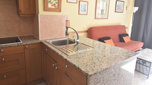 A kitchen with a granite countertop, a sink, and wooden cabinets. There are framed pictures on the wall and a red couch in the living area. - Apartment in Roses with pool, close to the beach and center, ideal for a comfortable and relaxing holiday. (Roses)