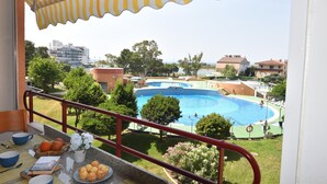 A balcony with a view of a swimming pool, a table with a book and a plate of food, and a railing.