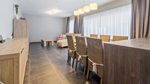 Ein modernes Wohnzimmer mit Sofa, Esstisch und Stühlen. Am linken Rand befindet sich ein kleiner Holzschrank und eine Pflanze.