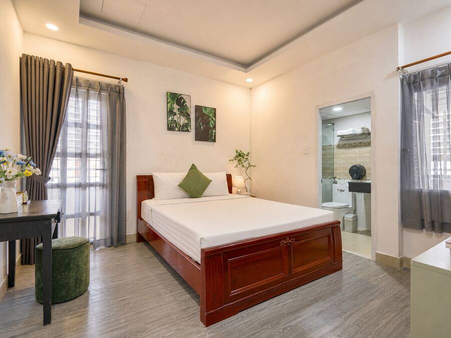 A hotel room with a wooden bed, a bedside table with a vase of flowers, a green ottoman, and a bathroom visible through a glass door.