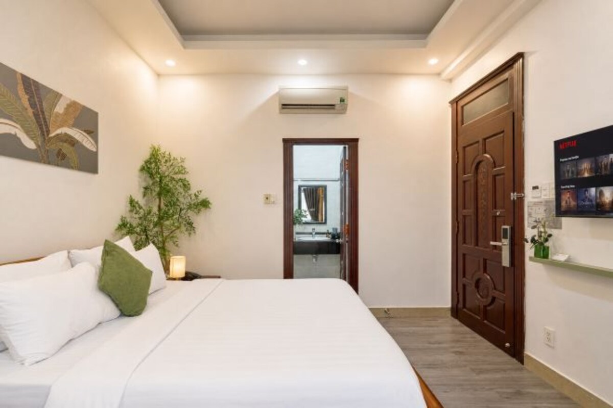 A hotel room with a bed, a nightstand, a potted plant, a door, and a wall-mounted shelf.
