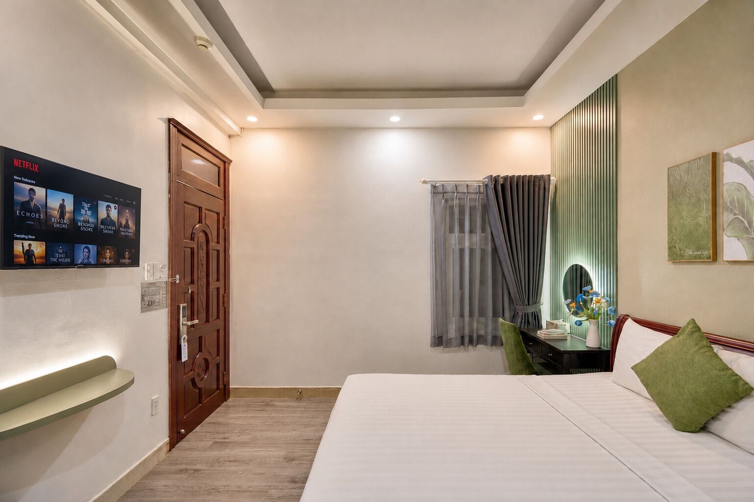 Habitación de hotel con cama, escritorio con silla, ventana con cortinas, televisor de pared mostrando contenido de Netflix y una puerta.