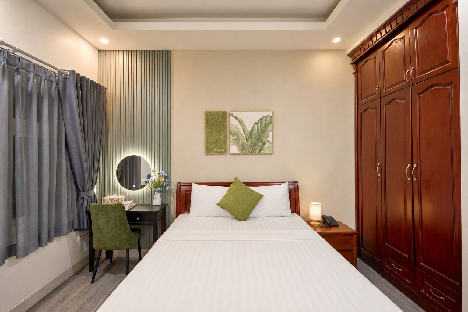 A hotel room with a bed, a desk with a chair, a mirror, a nightstand with a lamp, and a wardrobe.
