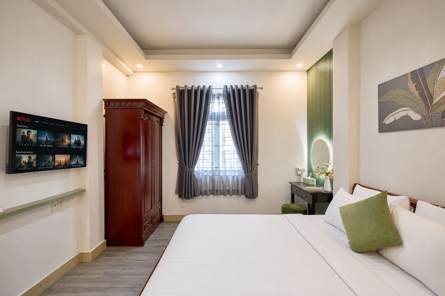 Habitación de hotel con una cama, televisión montada en la pared, armario, ventana con cortinas y un cuadro en la pared.