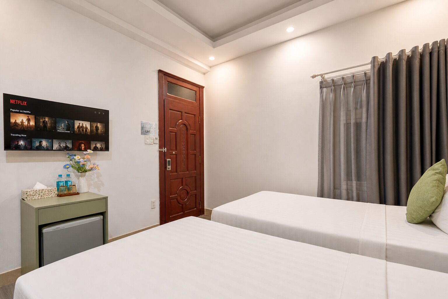 A hotel room with two beds, a wooden door, a wall-mounted TV displaying Netflix content, and a small table with a vase and bottles.