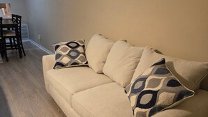 A beige sofa with patterned cushions in a living room with wooden flooring and a dining area with a table and chairs. - Tranquil 2-bedroom condo in Augusta with AC, WiFi (Augusta)