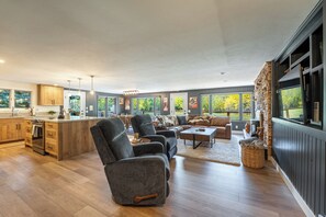 A spacious living area with a large sofa, a coffee table, and a fireplace. There is a kitchen area with wooden cabinets and a large island. The room has large windows offering views of trees.