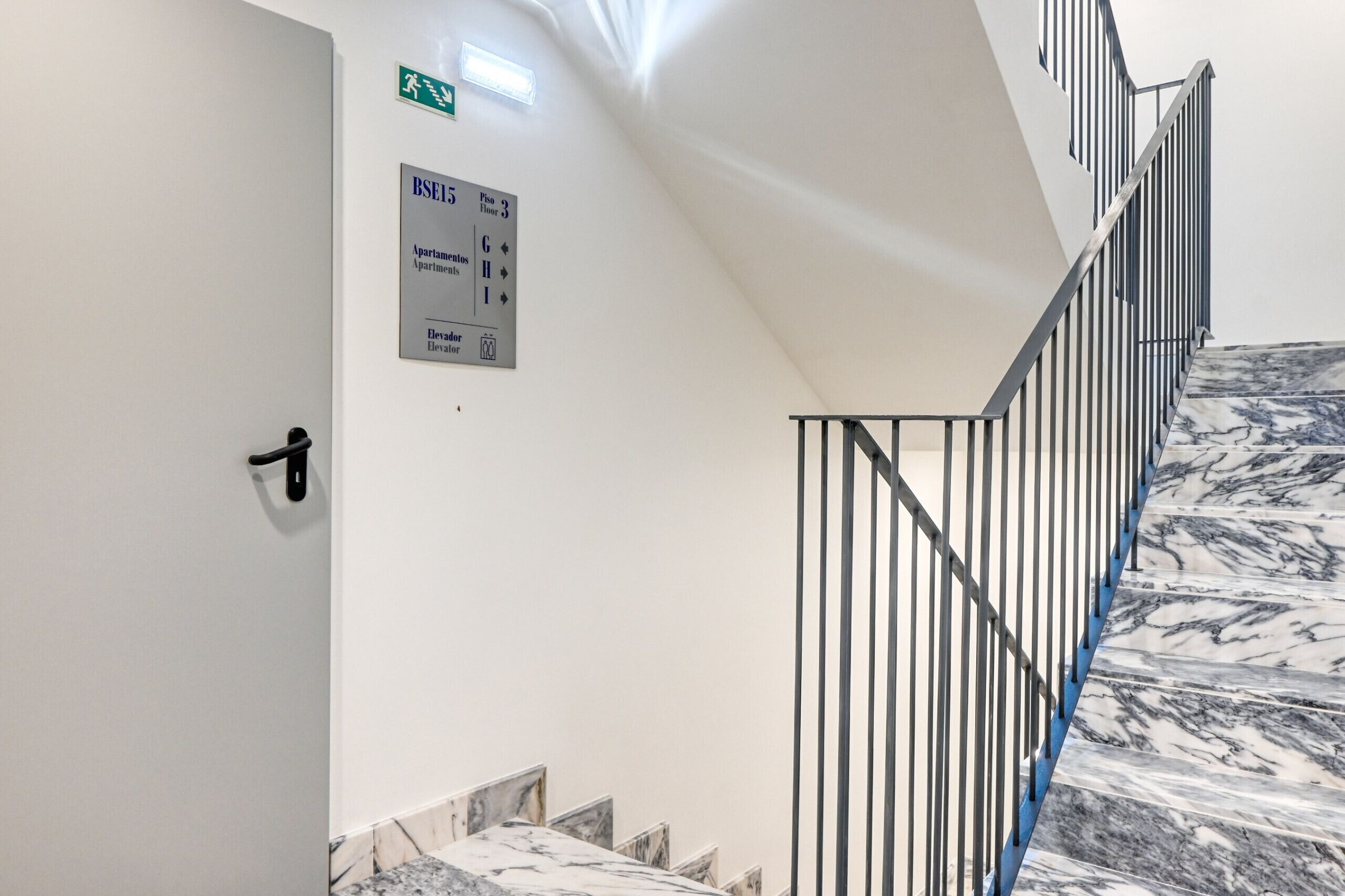 Eine Treppe mit Marmorstufen und Metallgeländern, eine Tür mit Griff und ein Schild, das BSE5 mit Wohnungsbezeichnungen und Stockwerken angibt.