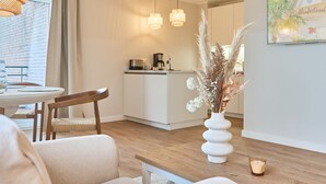 Una cocina moderna con armarios blancos y una cafetera, un comedor con una mesa y sillas de madera, y un jarrón con plantas secas en el suelo.