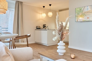 Une cuisine moderne avec des meubles blancs et une machine à café, un coin repas avec une table et des chaises en bois, et un vase contenant des plantes séchées, posé sur le sol.