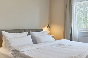 En pænt redt seng med hvide sengetøj og puder, en sengetlampe samt et vindue med gardiner.