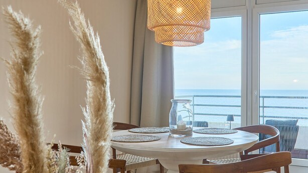 Una mesa redonda para comer con mantel blanco, rodeada de sillas de madera. Sobre la mesa, un tarro de vidrio con arena y una pequeña planta.