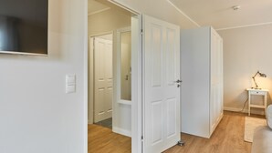 Una habitación con piso de madera, paredes blancas, una puerta blanca, una pequeña mesa blanca con una lámpara y un televisor montado en la pared.