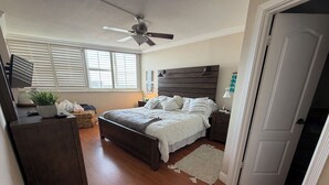 Ein Schlafzimmer mit Bett, Nachttischen, Deckenventilator, Fenster mit Jalousien, einer Tür und einem kleinen Teppich.