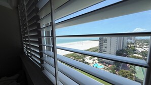 Blick aus einem Fenster mit Rollläden auf einen Strand, einen Pool und umliegende Gebäude.