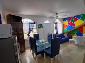 Ein Essbereich mit einem Glastisch und Stühlen, einem blauen Sofa, einem weißen Sessel und einer Wand mit farbenfrohen geometrischen Mustern.
