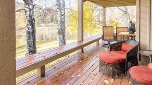 A covered outdoor seating area with wooden benches and wicker chairs, overlooking a yard with trees and a building in the background. - Elkhorn Retreat: Condo with Pond Views, Pool & Pet-Friendly (Sun Valley)