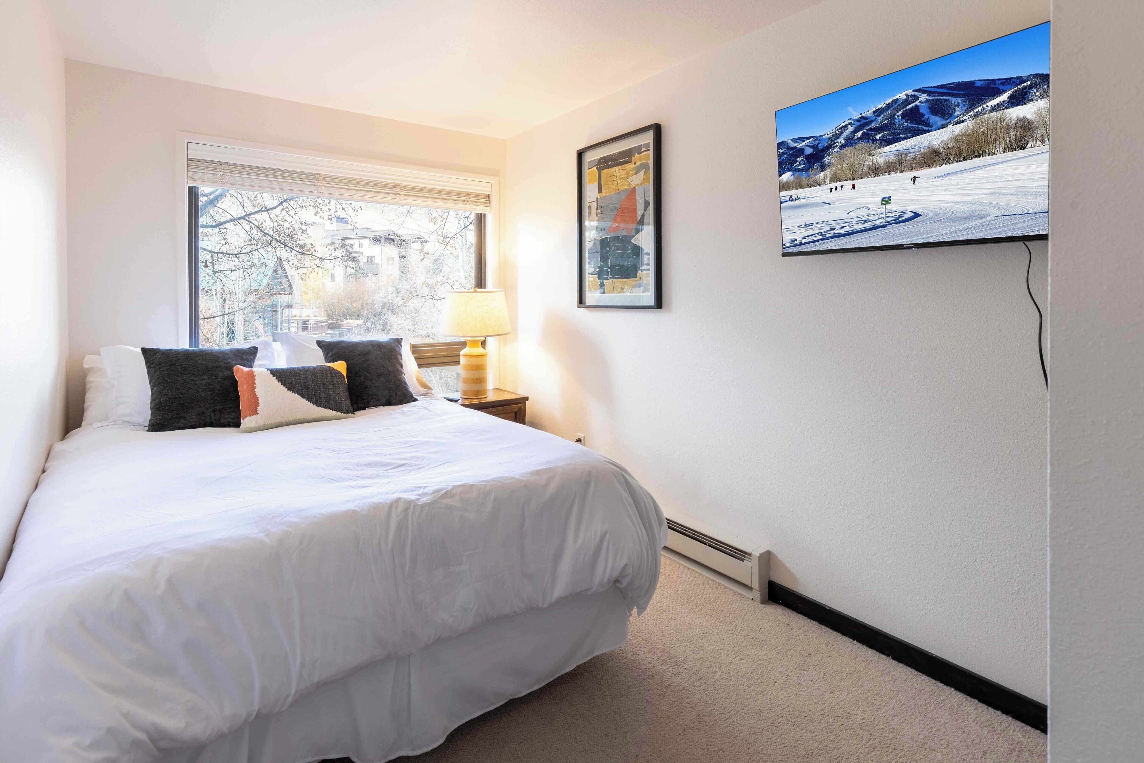 Une chambre d’hôtel avec un grand lit, une fenêtre avec des stores, un téléviseur fixé au mur et un tableau encadré.