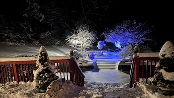 A snow-covered outdoor area with a wooden deck, snow-covered trees, and a pathway leading to a lit area with blue lights.