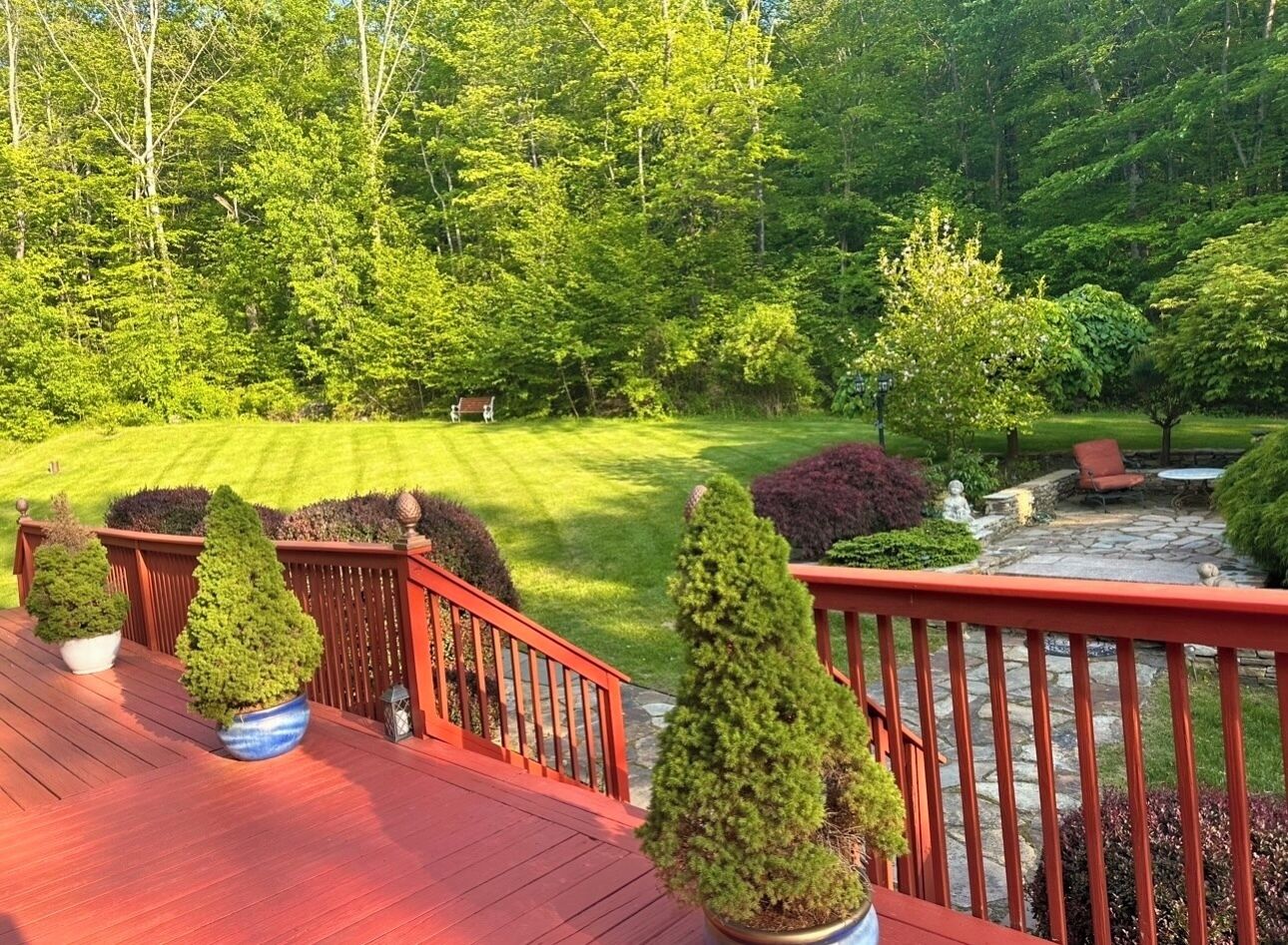 A wooden deck with potted coniferous trees, a stone patio with outdoor furniture, and a well-maintained lawn with a variety of shrubs and trees in the background.