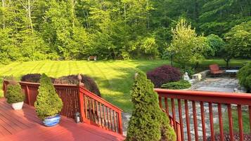 A wooden deck with potted coniferous trees, a stone patio with outdoor furniture, and a well-maintained lawn with a variety of shrubs and trees in the background.