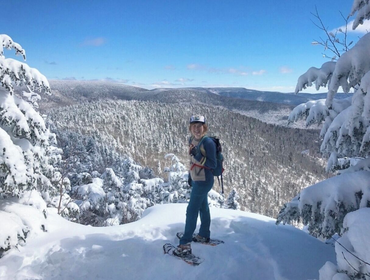Una persona de pie en una montaña nevada con una mochila, rodeada de árboles cubiertos de nieve y un cielo despejado.