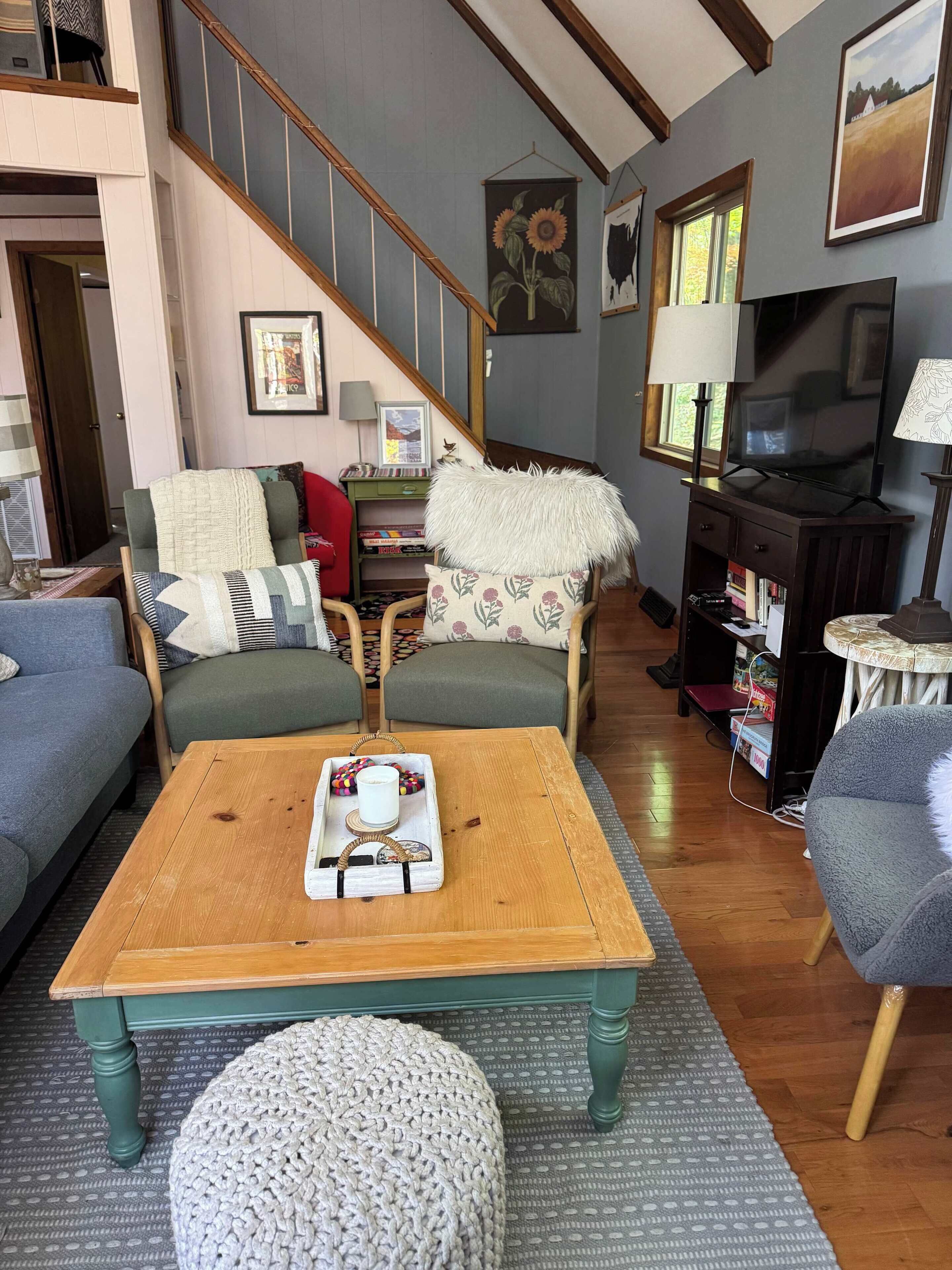 A cozy living room with a wooden coffee table, a sofa, and a chair. There is a staircase in the background, and a television on a stand.