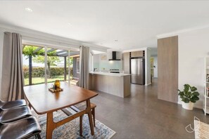 A modern kitchen with a central island, a dining area with a wooden table and chairs, and a view of a garden through large sliding doors.