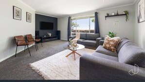 Una sala de estar moderna con un sofá gris, una mesa de centro y un televisor. Hay dos sillones, una alfombra y una ventana con cortinas.