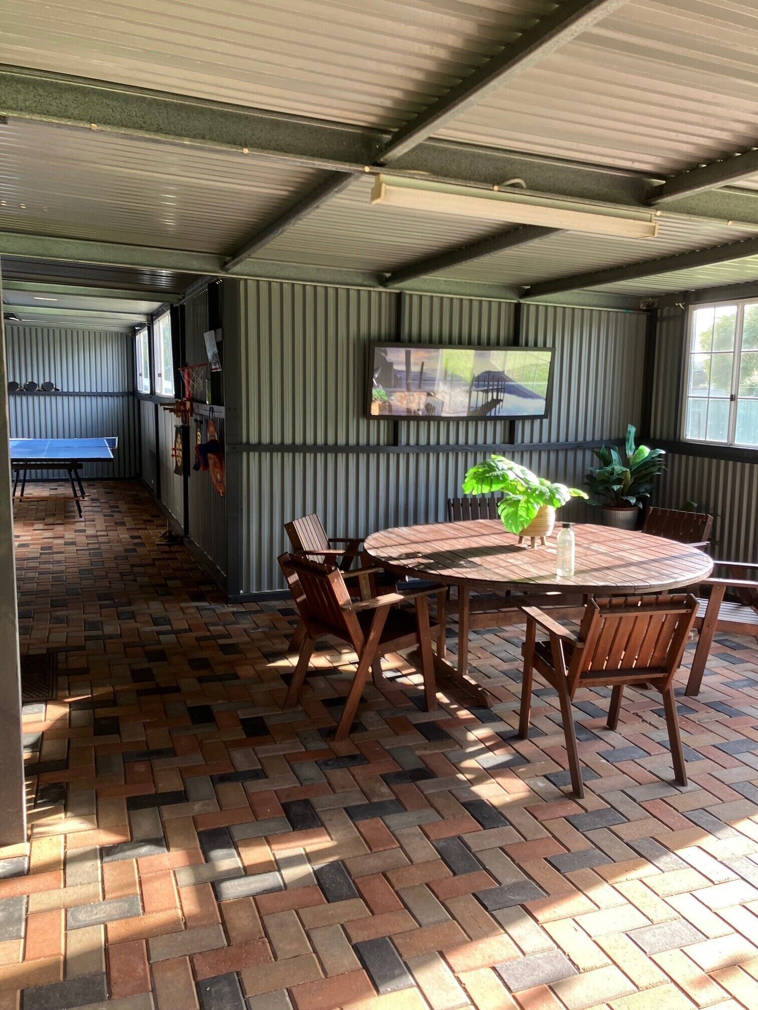 Un patio couvert avec un sol en briques, une table ronde en bois entourée de chaises, et une table de ping-pong à l’arrière-plan.