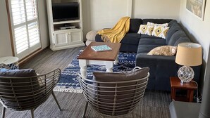 Uma sala de estar com um sofá cinza escuro, duas cadeiras pretas com estrutura de arame, uma mesa de centro, uma TV sobre um móvel branco, um tapete bege e uma janela com persianas.