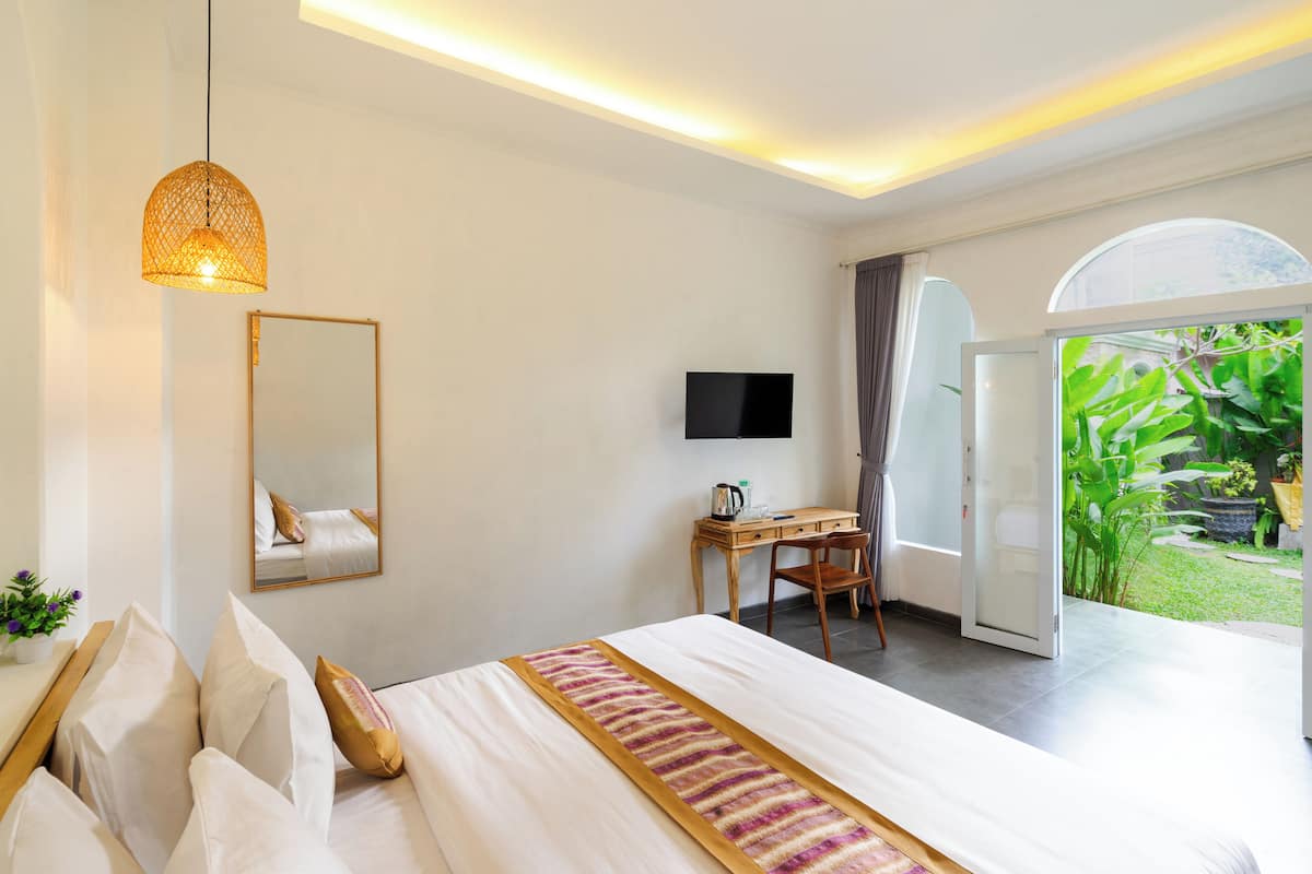 A modern bedroom with a large bed, a desk, a television, and a view of a garden through an open door.
