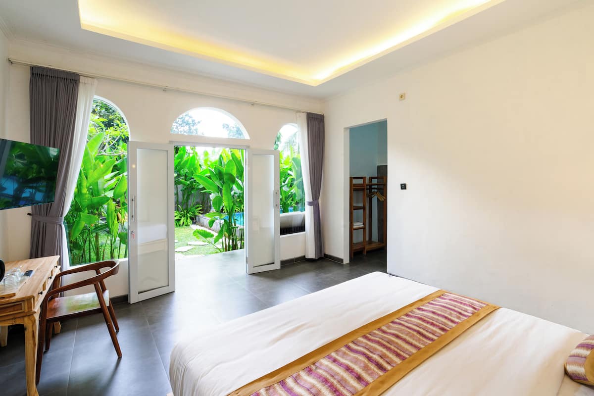 A hotel room with a large bed, a desk with a chair, a TV, and a view of lush greenery through open doors.