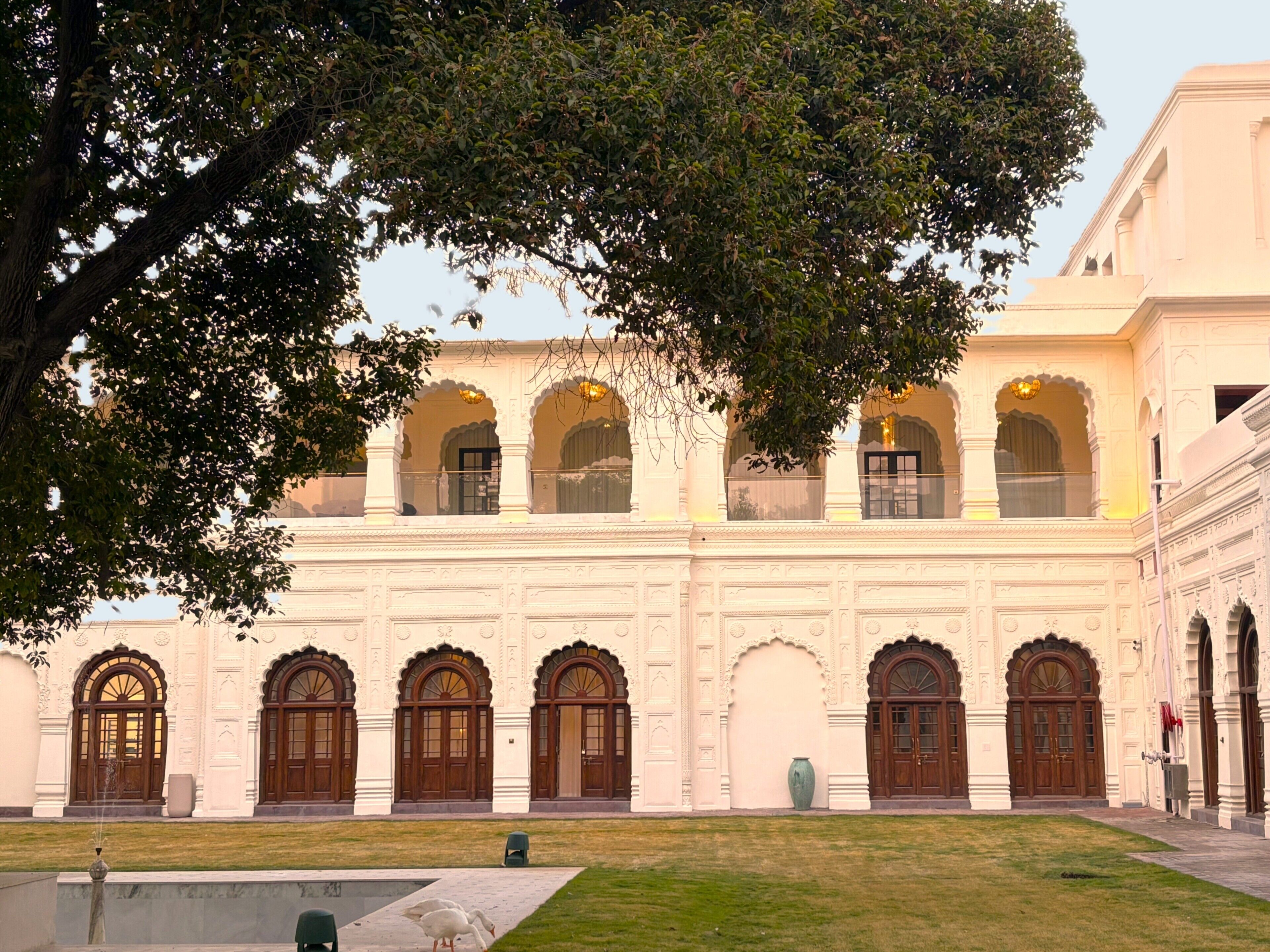 Un edificio con múltiples ventanales arqueados y puertas, un patio con una fuente y un árbol grande al frente.
